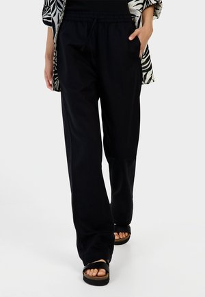 Zwarte, loszittende broek met een elastische tailleband en trekkoord, gemaakt van lichte stof. Gecombineerd met zwarte platte sandalen.