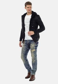 Zwarte gebreide zip-up hoodie met een textuurpatroon, gedragen over een witte graphic T-shirt. Lichtblauwe versleten jeans en bruine veterschoenen.