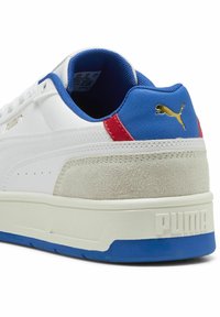 Sneaker sintetico bianco con accenti blu e rossi. Presenta un pannello in suede grigio testurizzato e una suola bianca robusta con logo impresso.