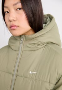Alyvuogių žalios spalvos pūkų striukė su aukštu apykaklės, priekyje užtrauktuku ir baltu Nike logo ant krūtinės. Lygus, lengvas audinys su kvadrato raštu.