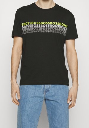 T-shirt noir à manches courtes avec texte "BOSS" répété horizontalement en blanc et jaune néon sur la poitrine, porté avec un jean bleu.