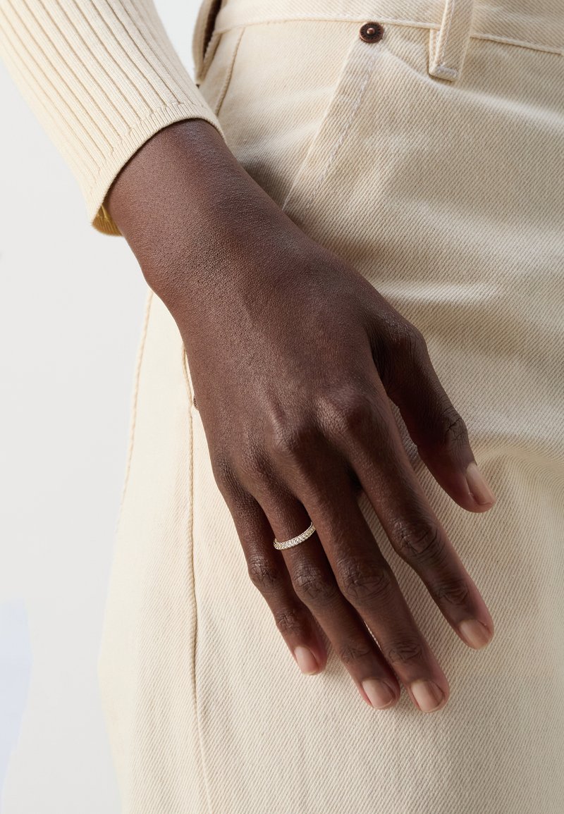Bague en argent avec des pierres transparentes présentée sur une main contre un fond de tissu beige avec une manche crème texturée et côtelée et un pantalon tan.
