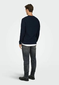 Pull en maille texturée bleu marine avec un col rond, associé à un jean gris foncé et des chaussures noires ; vu de dos.