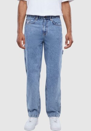 Mies, jolla on vaaleansiniset suorat farkut, joissa on sivutasku, valkoiset lenkkarit ja valkoinen t-paita, joka on työnnetty housuihin, yksivärisellä taustalla.