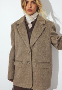 Femme portant un manteau marron texturé oversize par-dessus un col roulé blanc et une chemise foncée, debout devant un fond blanc uni.