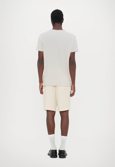 Camiseta gris claro, diseño liso; pantalones cortos crema con un patrón sutil; calcetines blancos; zapatos negros; visto desde atrás sobre un fondo blanco.