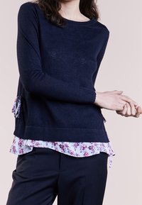 Marinblå stickad topp med långa ärmar, som har en blommig volang i kanten och sidodetaljer, kombinerad med mörka byxor.