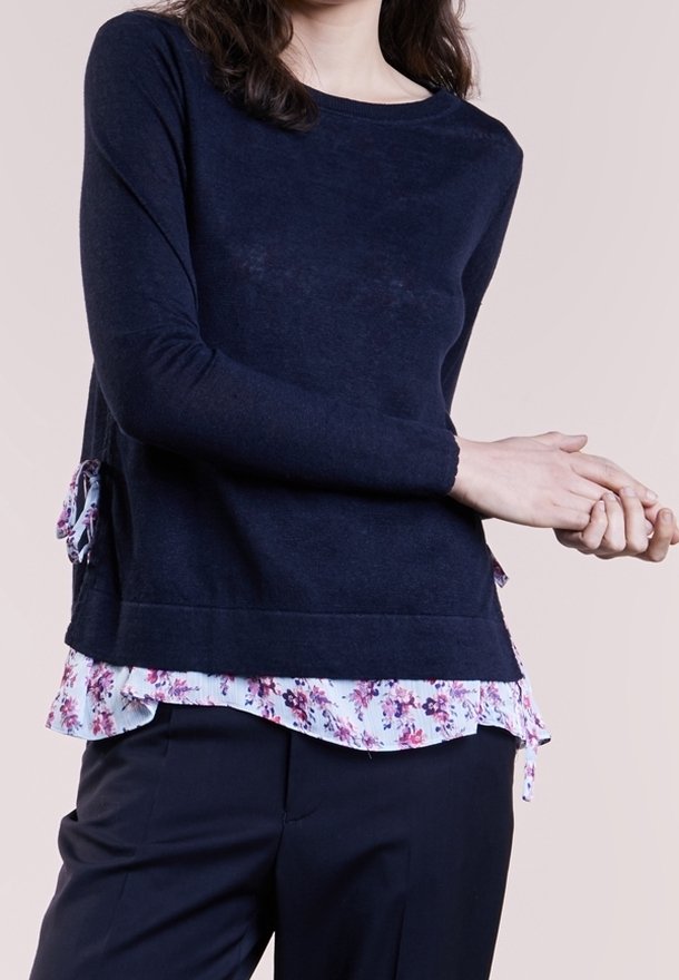 Marinblå stickad topp med långa ärmar, som har en blommig volang i kanten och sidodetaljer, kombinerad med mörka byxor.