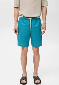 Pantaloni shorts turchesi realizzati in tessuto leggero, con una vita elasticizzata e un laccio beige, dal taglio rilassato, abbinati a sandali neri.