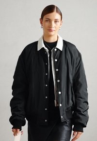 Veste bombers noire avec un col blanc, des boutons pression et des poignets côtelés. Texture lisse ; portée par-dessus un haut noir avec une jupe en cuir.