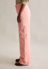 Spodnie szerokie z sztruksu w jasnoróżowym kolorze, z wysokim stanem, bocznymi kieszeniami i prążkowaną fakturą. Noszone z ciemnobrązowymi butami.