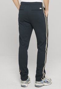 Marineblaue Jogginghose mit beigen Seitenstreifen, elastischem Bund und zwei Gesäßtaschen. Kombiniert mit grauen und blauen Sportschuhen.