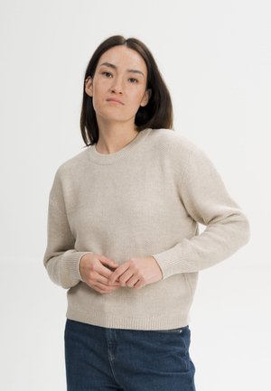 Vrouw met stijl donker haar, gekleed in een beige gebreide trui en blauwe spijkerbroek, staande tegen een effen lichte achtergrond.