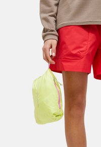 Pochette vert clair avec une fermeture éclair rose, fabriquée en matériau lisse, tenue dans une main, à côté de shorts rouges et d'une chemise à manches longues marron texturée.