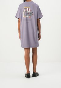 Světle fialové oversize tričkové šaty s krátkým rukávem. Ilustrace obchodu se stromy vzadu. Měkká bavlněná textura.