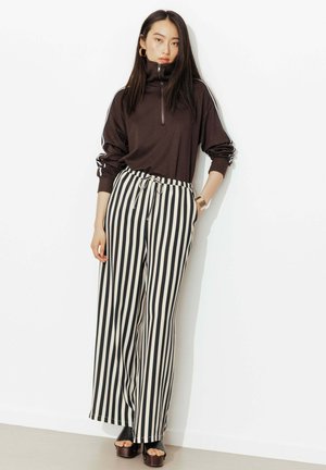 Femme portant un haut noir zippé à manches longues, un pantalon large à rayures verticales noir et blanc, et des chaussures compensées noires à bout ouvert, debout contre un mur blanc.