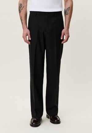 Uomo con pantaloni neri su misura, camicia bianca, scarpe in pelle scura e tatuaggi visibili sulle braccia, in piedi davanti a uno sfondo neutro.
