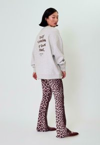 Šedá oversized mikina s hnědým textem na zadní straně, doplněná růžovými a hnědými kalhotami s leopardím vzorem a rozšířenými nohavicemi. Modelka stojí na světlejším pozadí.