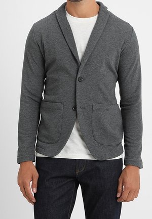 Mężczyzna w dopasowanej ciemnoszarej marynarce na białej koszulce i ciemnych dżinsach, stojący z ramionami swobodnie opuszczonymi wzdłuż boków.