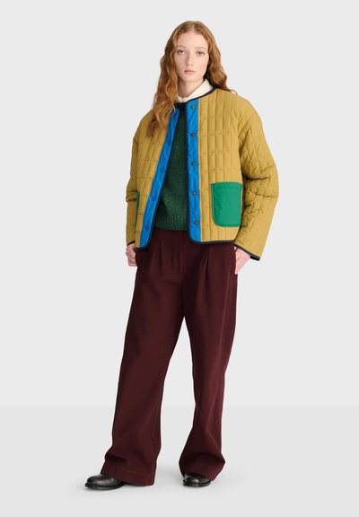 Jeune personne aux longs cheveux ondulés porte une veste matelassée moutarde avec bordure bleue, un pull vert, un pantalon large bordeaux et des chaussures noires sur fond blanc.