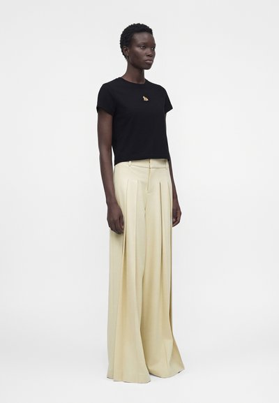 Γυναίκα φοράει μαύρο μπλουζάκι με κοντό μανίκι με μικρό έμβλημα αλεπούς και μπεζ παντελόνια με φαρδιά μπατζάκια και ψηλή μέση, στέκεται μπροστά σε λευκό φόντο.