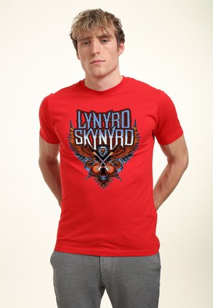 Jeune homme portant un T-shirt rouge Lynyrd Skynyrd avec un aigle et des guitares croisées, debout devant un fond uni.