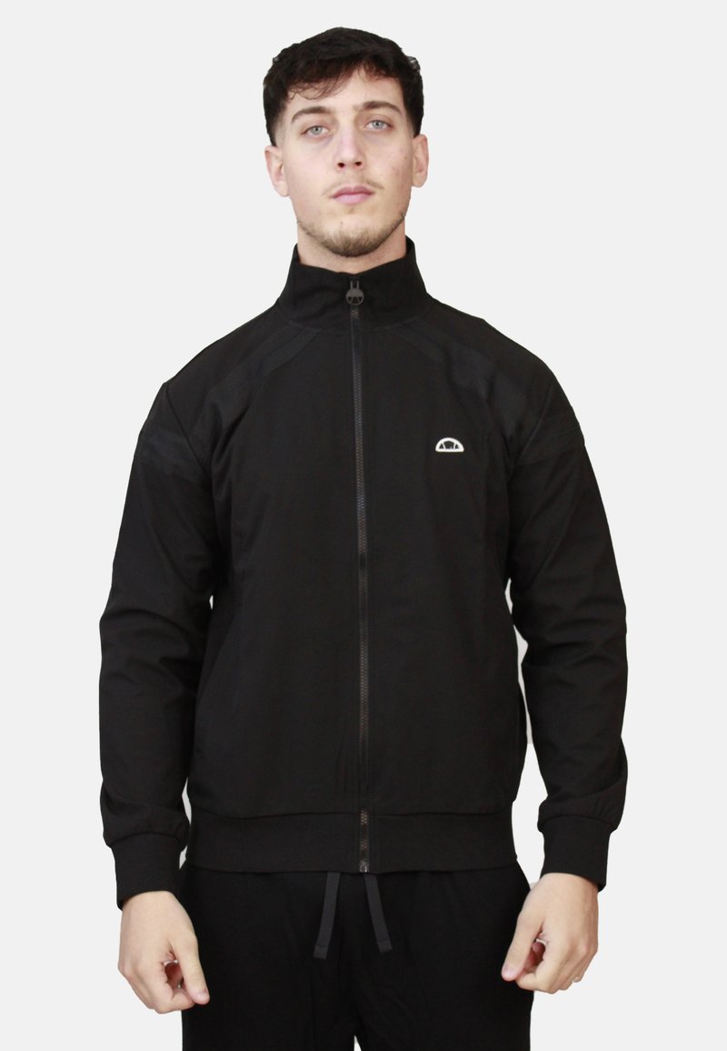 Giacca sportiva nera realizzata in tessuto liscio, dotata di colletto alto, zip a tutta lunghezza e un logo sul petto. Design aderente con cuciture ben definite.