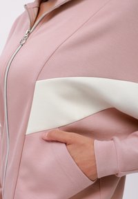 Růžová zipová mikina s bílým akcentním panelem, měkkou texturou látky, bočními kapsami a žebrovanými manžetami, zdůrazňující sportovní design.