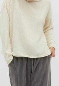 Cremfarbener Strickpullover mit lockerer Passform und gerippter Textur, kombiniert mit grauen schmal gestreiften Hosen mit Kordelzug.