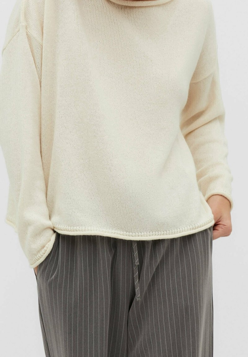 Cremfarbener Strickpullover mit lockerer Passform und gerippter Textur, kombiniert mit grauen schmal gestreiften Hosen mit Kordelzug.