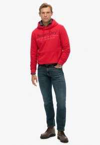 Czerwona bawełniana bluza z kapturem, z przodem kieszenią i sznurkiem, ozdobiona logo. Połączona z ciemnoniebieskimi dżinsami i brązowymi skórzanymi butami.
