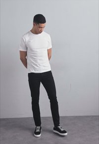 Bílý bavlněný tričko, černé slim-fit kalhoty a černé tenisky s bílou podrážkou. Osoba stojí s rukama za zády a hlavou mírně skloněnou.