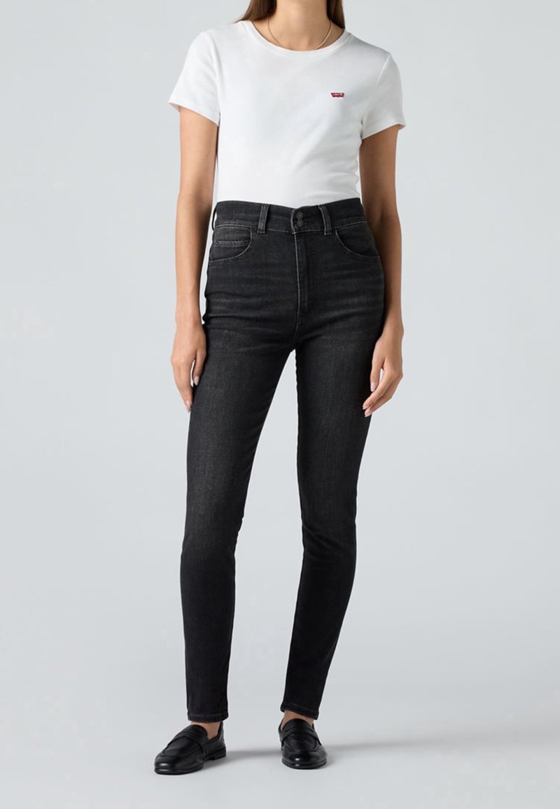 Femme portant un t-shirt blanc à manches courtes ajusté avec un petit logo rouge et un jean skinny noir taille haute avec des mocassins noirs.