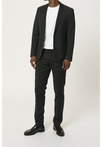 Completo nero con giacca a un petto, pantaloni affusolati e una maglietta bianca sotto. Scarpe in pelle nera lucida completano il look.
