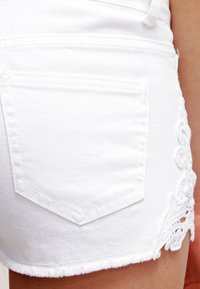 Primer plano de unos pantalones cortos de mezclilla blanca con dobladillo deshilachado, bolsillo trasero y detalle de encaje floral en el lado, llevados en la cadera y el muslo de una persona.