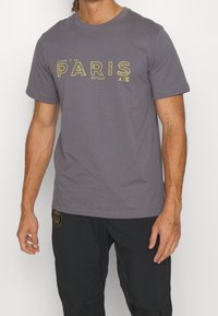 Muž v šedém triku s krátkým rukávem s žlutým nápisem "PARIS" a černých kalhotách se žlutým logem Paris Saint-Germain na levém stehně.