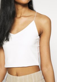 Crop top blanc côtelé avec fines bretelles spaghetti, présentant un décolleté en V et une coupe ajustée. Texture lisse, porté avec un pantalon taille haute.