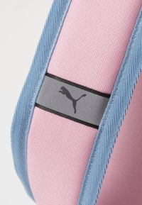 Mochila Puma en materiales rosa y azul con una superficie texturizada; cuenta con una correa gris con un logo negro y costuras en contraste.