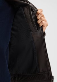 Dunkelbraune Lederjacke von innen, die ein glattes schwarzes Futter und eine Seitentasche zeigt. Der Stoff hat einen leichten Glanz und eine weiche Textur.