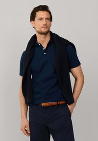 Muž v tmavomodrej polo košeli a nohaviciach, s čiernym svetrom prehodeným cez ramená, stojaci pred jednoliatou sivou stenou.