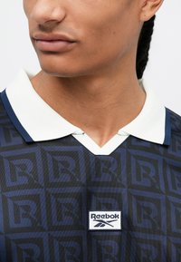 Polo de culoare albastru închis, cu un model texturat în diamante, guler alb și patch cu logo-ul Reebok pe față, având un design cu decolteu în V.
