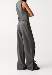 Haut sans manches gris sur mesure avec un nœud au dos, associé à un pantalon gris à jambes larges comprenant des fentes latérales et des poches arrière.