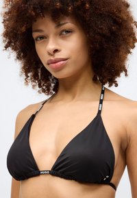 Μαύρο μπικίνι τοπ με τρίγωνες cups, συγκεντρωμένο κέντρο και λεπτές ράντες που φέρουν λογότυπο. Λείο υφαντό και ρυθμιζόμενα σχεδιαστικά στοιχεία.
