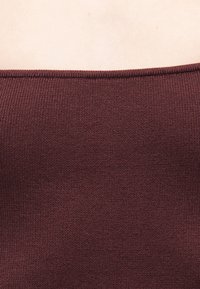 Κοντινό πλάνο ενός ατόμου που φοράει μπορντό ριμπ πλεκτό τοπ με ευθεία γραμμή λαιμού, σε ουδέτερο φόντο.