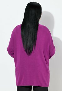 Person mit langen schwarzen Haaren, die einen locker sitzenden lila Pullover trägt, steht vor einer weißen Wand und schaut weg.