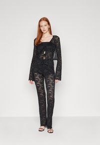 Svart spetsset med långärmade blusar och byxor, med scallopkanter och en knytdetalj. Använt tillsammans med en liten svart handväska och sandaler med remmar.