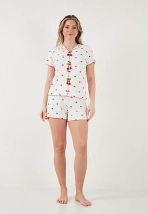 Frau steht barfuß und trägt einen weißen kurzärmligen Pyjama mit kleinen Kirschmotiven und roten Kirschknöpfen am Hemd.