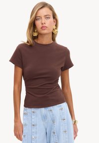 Femme aux cheveux blonds portant un haut marron ajusté à manches courtes, un pantalon taille haute bleu clair avec des boutons argentés, des boucles d'oreilles dorées et une montre-bracelet.