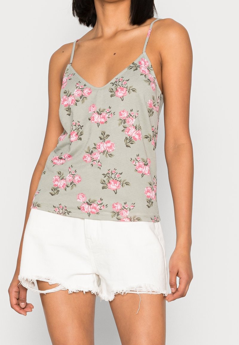 Débardeur fleuri avec un col en V, des bretelles fines et un fond gris clair orné de roses roses. Associé à un short blanc déchiré.