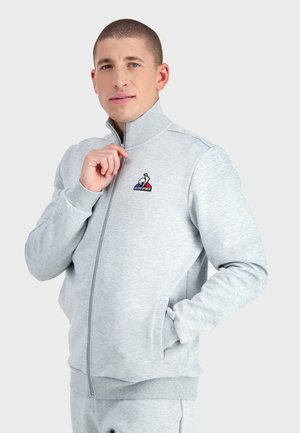 Chaqueta gris con cremallera hecha de tejido suave, con cuello alto, puños de canalé y un logo en el lado izquierdo del pecho. Textura suave, corte casual.